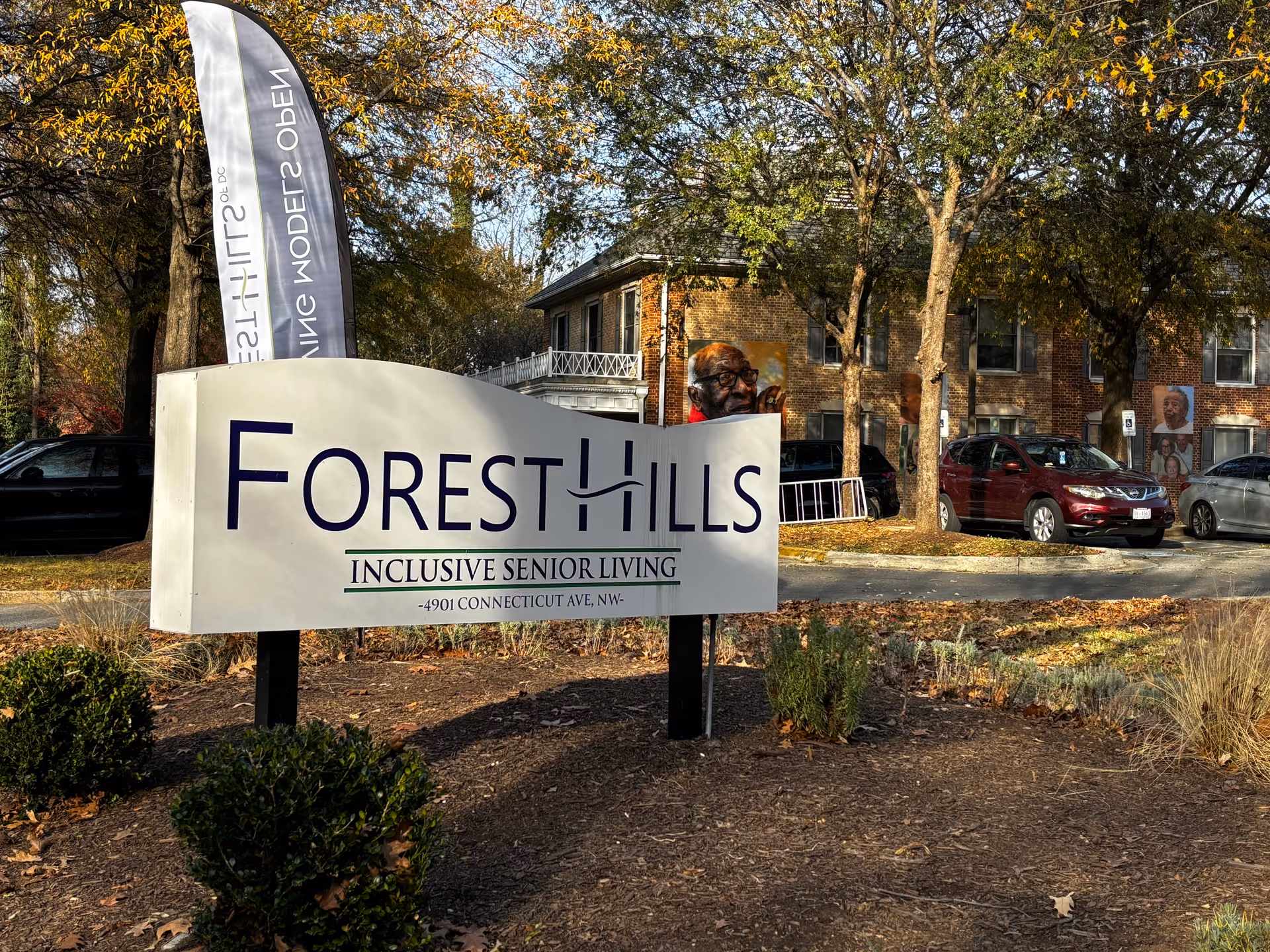 Outdoor view of the entrance sign for Forest Hills Inclusive Senior Living at 4901 Connecticut Ave NW, with trees, parked cars, and a brick building in the background.