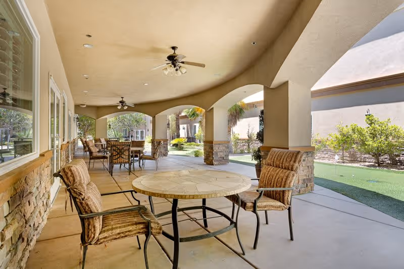Covered outdoor patio area with round tables and cushioned chairs, ceiling fans, stone pillars, and a view of a garden with greenery and palm trees.