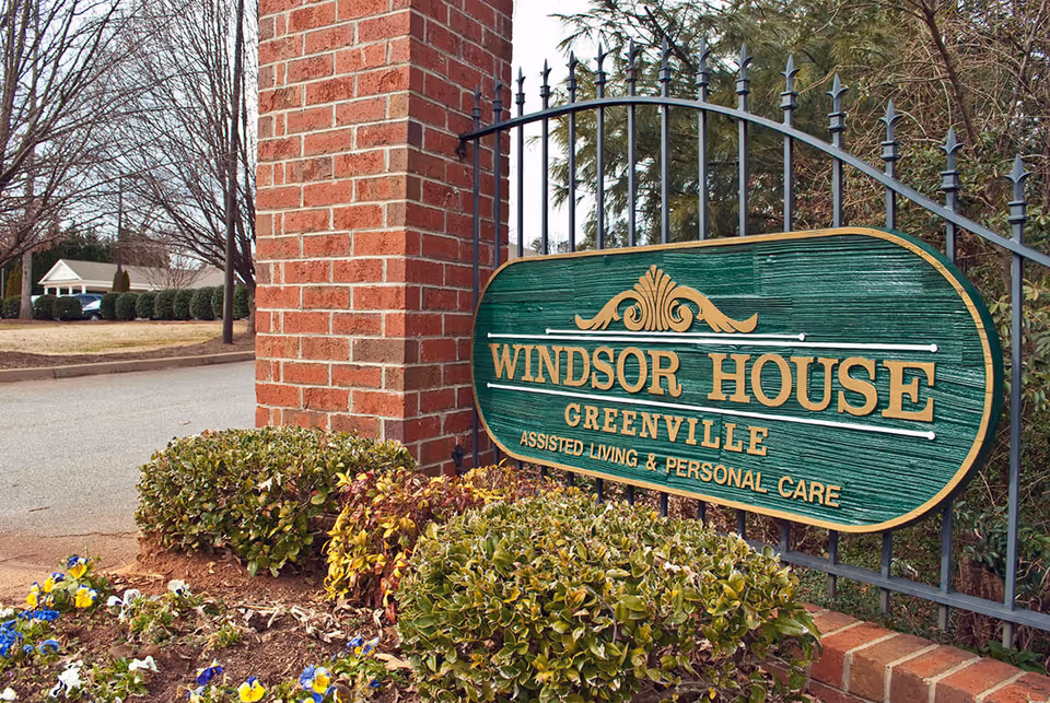 A green wooden sign with gold lettering that reads 'Windsor House Greenville Assisted Living & Personal Care' mounted on a black metal fence next to a brick pillar, surrounded by bushes and flowers with a road and trees in the background.