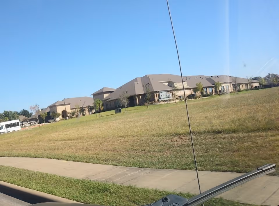 Exterior view of a large single-story brick building with multiple windows and a sloped roof, surrounded by a grassy lawn and a sidewalk in front. A white shuttle bus is parked on the left side near the building under a clear blue sky.