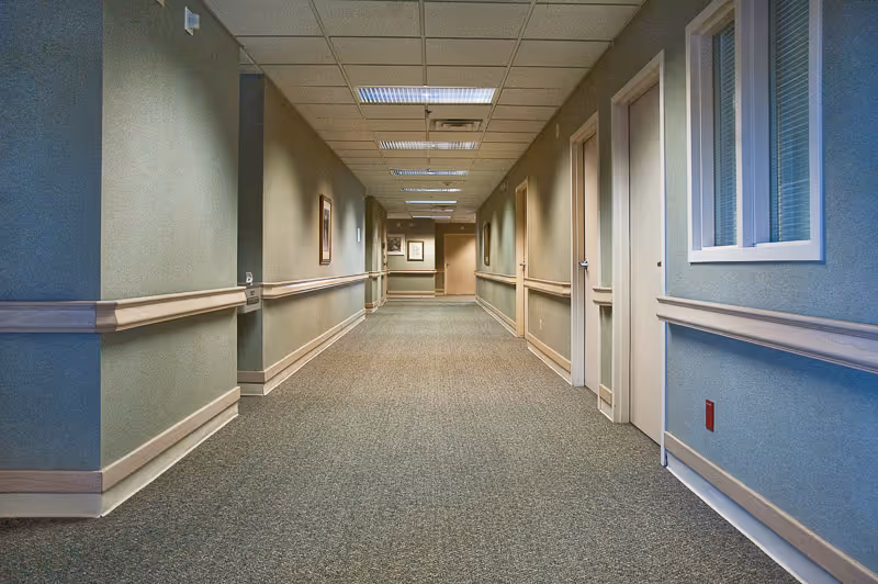 A long, empty hallway in a senior living facility with beige walls, carpeted floor, handrails on both sides, several closed doors, framed pictures on the walls, and ceiling lights.
