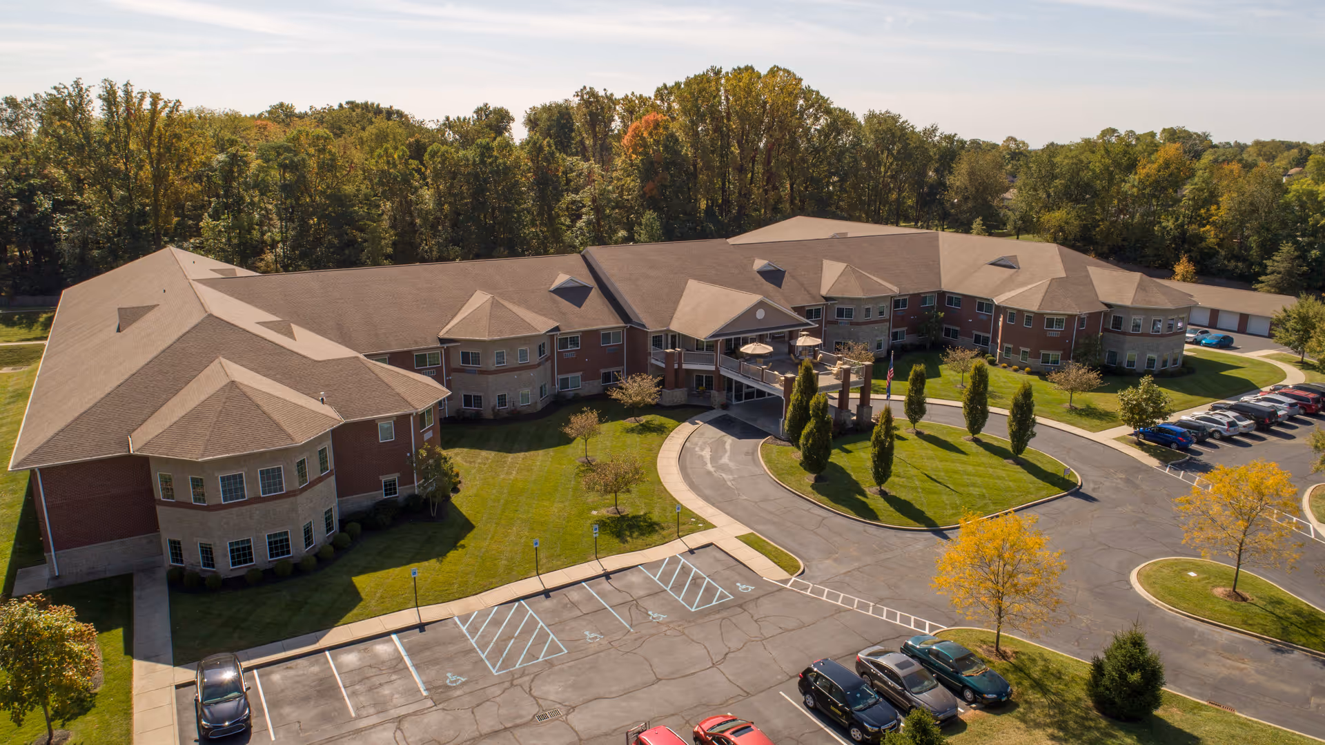 Aerial view of Magnolia Springs East Louisville, a large senior living facility with a U-shaped building surrounded by green lawns, trees, and a parking lot with several cars. The building has multiple windows and a covered entrance with a circular driveway.