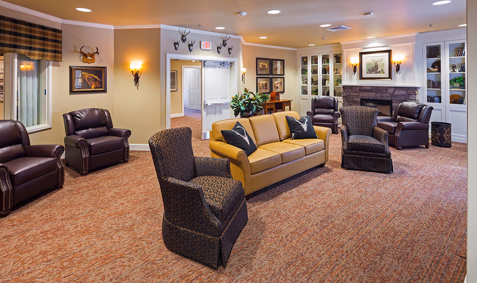 A cozy living room in a memory care community featuring a tan leather sofa with two black pillows, two patterned armchairs, and four dark brown leather armchairs arranged around a stone fireplace. The room has beige walls adorned with framed pictures and mounted deer heads. Built-in white shelves flank the fireplace, holding books and decorative items. The carpet is a warm, textured pattern, and soft lighting creates a welcoming atmosphere.