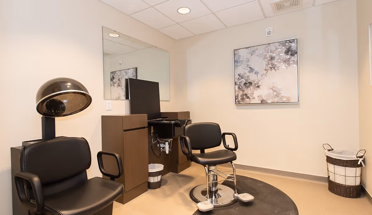 Interior of a hair salon area in an assisted living facility featuring two black salon chairs, a hair dryer chair, a large mirror on the wall, a small sink with cabinetry, a framed abstract floral painting, and a waste basket in the corner.