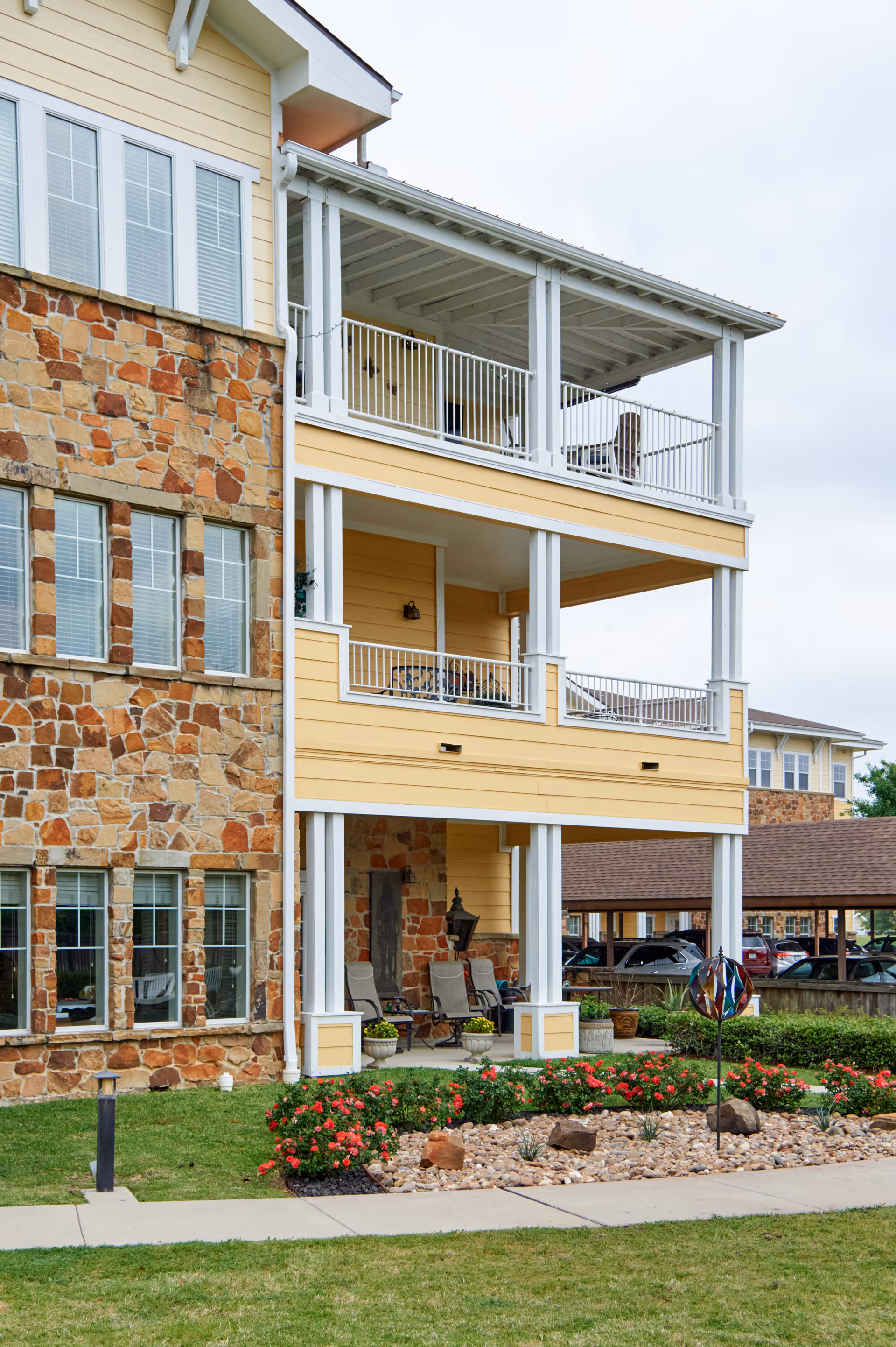 Exterior view of a senior living facility building with stone and yellow siding, featuring two covered balconies with white railings and outdoor seating. There is a landscaped garden with red flowers and rocks in front, along with a sidewalk and a parking area in the background.