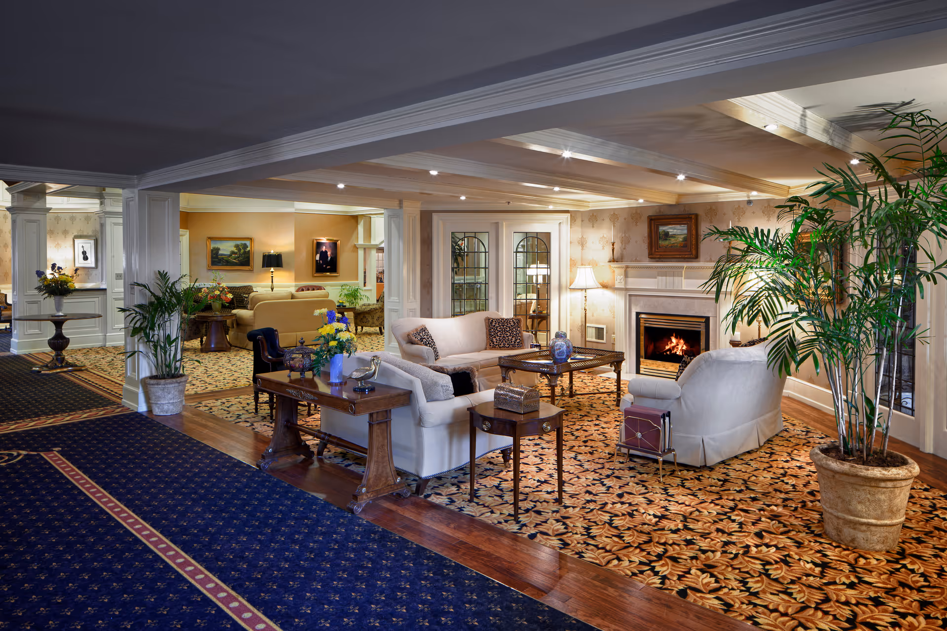 A spacious and elegantly decorated living room area in a senior living facility featuring comfortable white sofas, a wooden coffee table, a fireplace with a lit fire, large potted plants, framed paintings on the walls, and patterned carpet flooring. The room is well-lit with recessed ceiling lights and table lamps, creating a warm and inviting atmosphere.