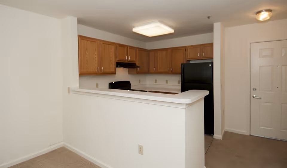 Small kitchenette with wood cabinets, a black refrigerator and stove behind a white breakfast bar and an entry door to the right.