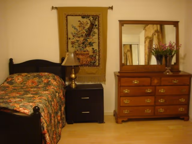 A bedroom with a single bed covered in a floral patterned bedspread, a black nightstand with a brass lamp, a wall hanging with a floral design, and a wooden dresser with a large mirror and a vase of flowers on top.