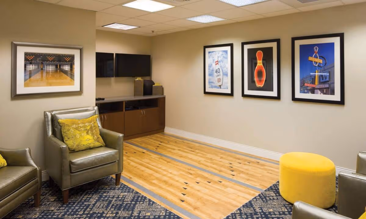 A cozy indoor recreational room with a small bowling lane, two leather armchairs with yellow patterned pillows, a yellow round ottoman, a wall-mounted flat screen TV, and framed pictures of bowling pins and signs on the walls.