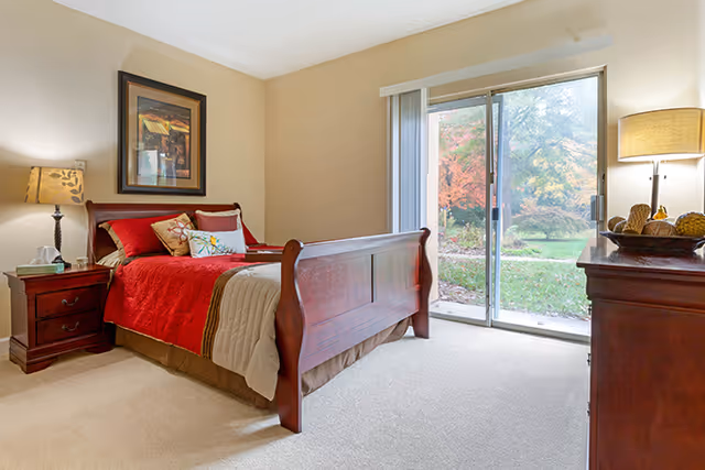 Sunny bedroom with a wooden sleigh bed dressed in red bedding, nightstand and dresser, and a sliding glass door overlooking a garden.