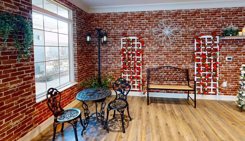 A cozy indoor seating area with exposed brick walls, a large window, two black ornate metal chairs with a matching round table, a wooden bench with a metal backrest, decorative white trellises adorned with red flowers, a black vintage-style street lamp, and a wooden floor.