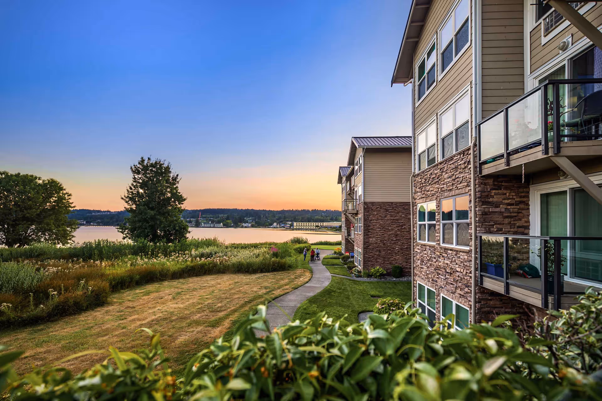 View of a senior living facility named Crista Shores during sunset, showing a multi-story building with balconies on the right, a paved walkway leading through green landscaped grounds, trees, and a body of water in the background under a clear sky.