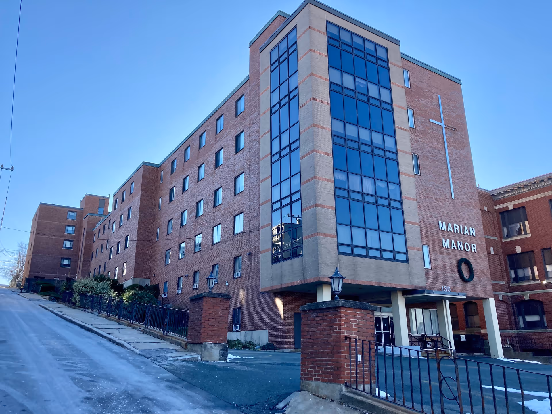 Exterior view of Marian Manor Nursing Home, a multi-story brick building with large vertical windows and a cross mounted on the front facade. The entrance is visible with a covered drop-off area and brick pillars with lanterns. The sky is clear and blue.