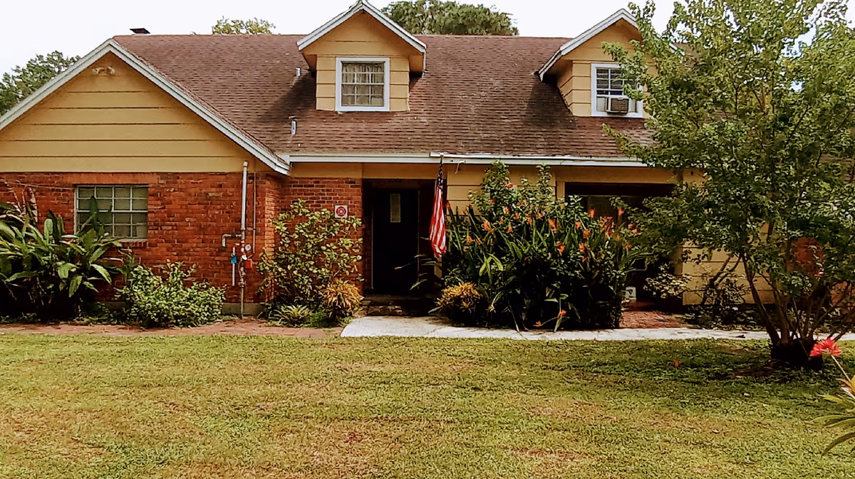Front exterior view of a single-story residential building with a brick and yellow siding facade, two dormer windows on the roof, an American flag near the entrance, and a well-maintained lawn with various shrubs and trees.