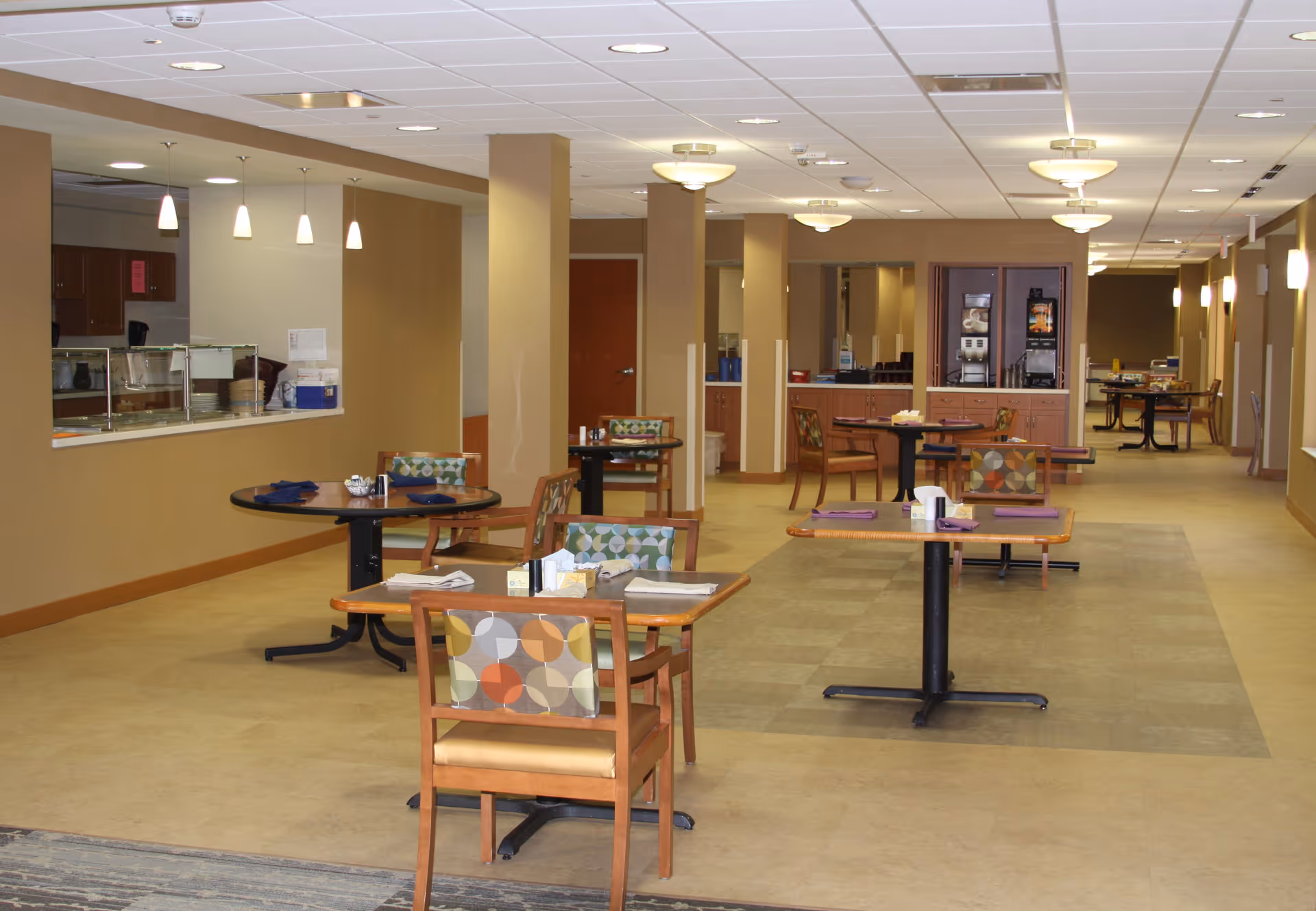 Interior view of a dining area in a senior living facility with several tables and chairs arranged neatly. The tables have placemats, napkins, and condiments. The room has beige walls, ceiling lights, and a serving counter on the left side. In the background, there is a beverage station and more seating.
