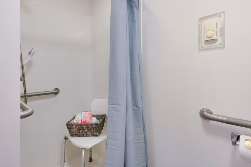 Accessible shower stall with a blue curtain, wall grab bars, a shower chair holding a basket of toiletries, and a handheld showerhead.