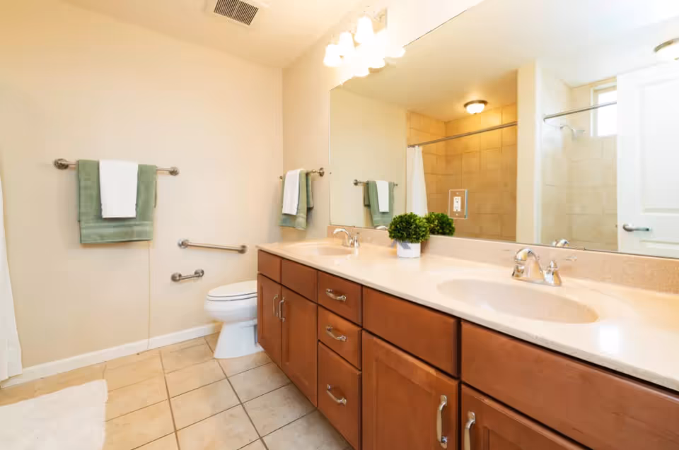 A clean and well-lit bathroom featuring a double sink vanity with wooden cabinets, a large mirror above the sinks, a toilet with grab bars on the wall, a towel rack with green and white towels, and a tiled shower area with a curtain and a small window.