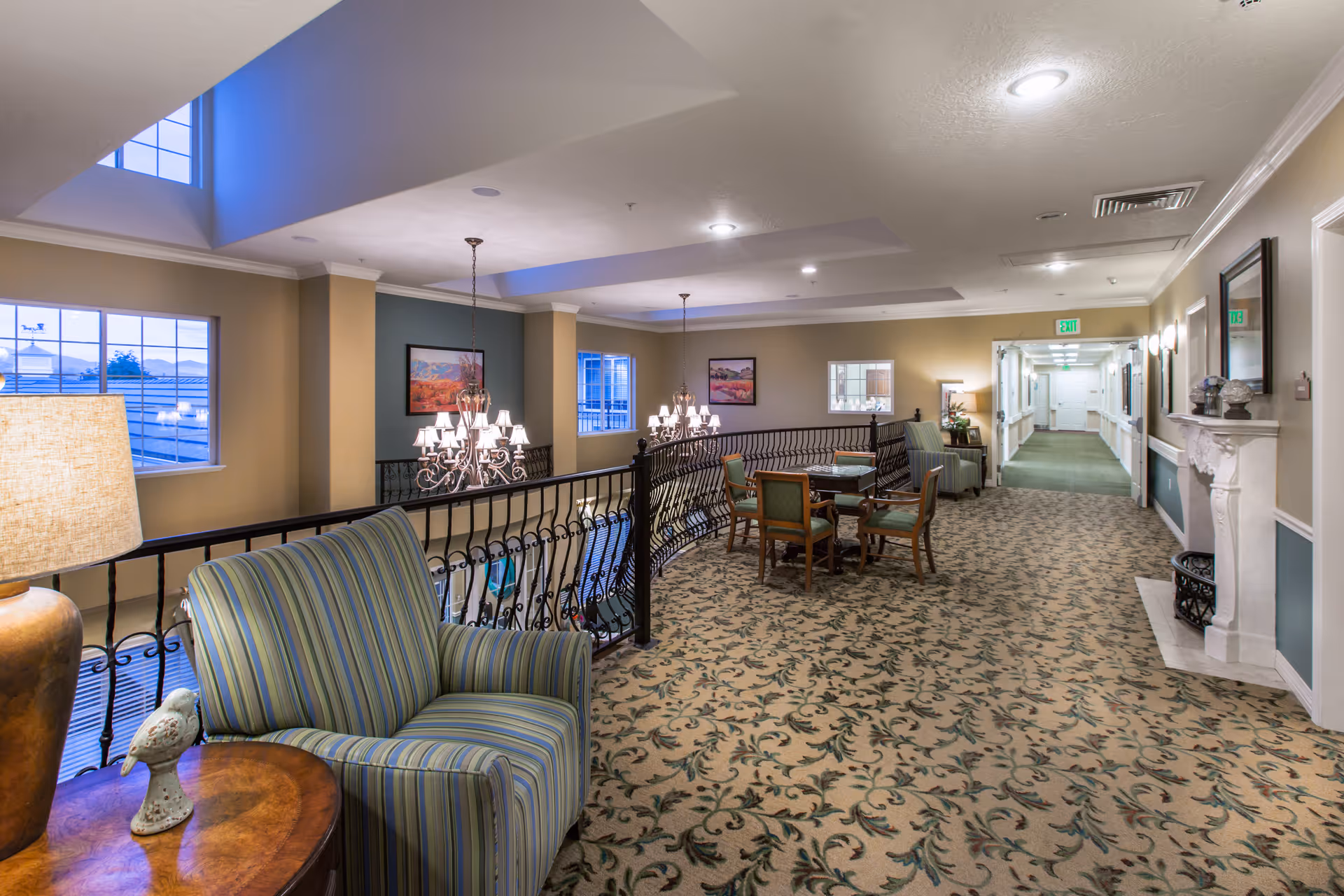 A spacious and well-lit interior common area with patterned carpet, striped armchairs, a wooden side table with a lamp and decorative bird, a table with four chairs, chandeliers hanging from the ceiling, framed artwork on the walls, and a hallway leading to other rooms.