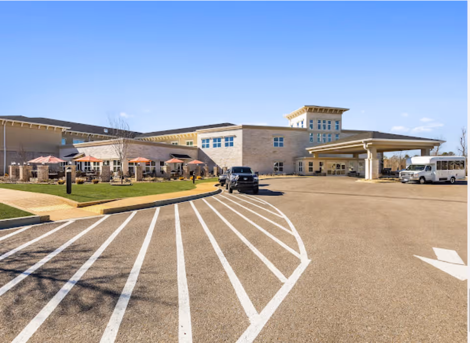 Exterior view of a senior living facility with a large parking area in front. The building has a covered entrance, multiple windows, and an outdoor patio area with tables and umbrellas. Several vehicles, including a black truck and a white shuttle bus, are parked near the entrance. The sky is clear and blue.