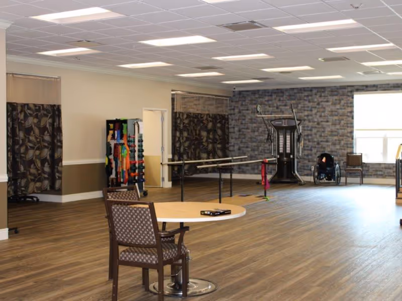 A spacious rehabilitation room with wooden flooring and a patterned accent wall. The room contains parallel bars for walking exercises, a multi-functional exercise machine, a wheelchair, a rack with various exercise bands and weights, and a round table with a chair in the foreground. Large windows allow natural light to brighten the space.