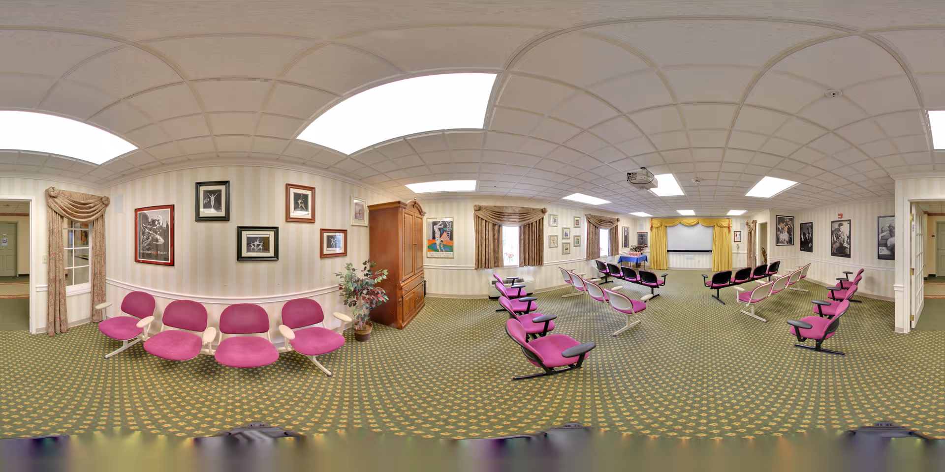 A large meeting or activity room with rows of purple cushioned chairs arranged facing a stage area with a projector screen and yellow curtains. The room has patterned green carpet, striped wallpaper, framed pictures on the walls, and several windows with draped curtains. There is a wooden cabinet and a potted plant near the entrance.