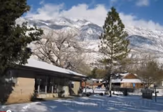Snow-covered outdoor area of a residential facility with a single-story building, trees, and mountains in the background under a partly cloudy sky.