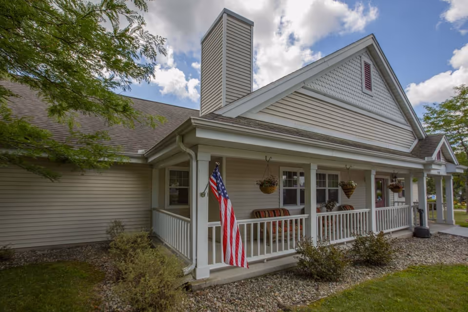 Exterior view of a single-story building with beige siding and a covered porch. The porch has white railings, hanging flower baskets, and cushioned chairs. An American flag is displayed on one of the porch columns. The building is surrounded by a lawn, bushes, and trees under a partly cloudy sky.