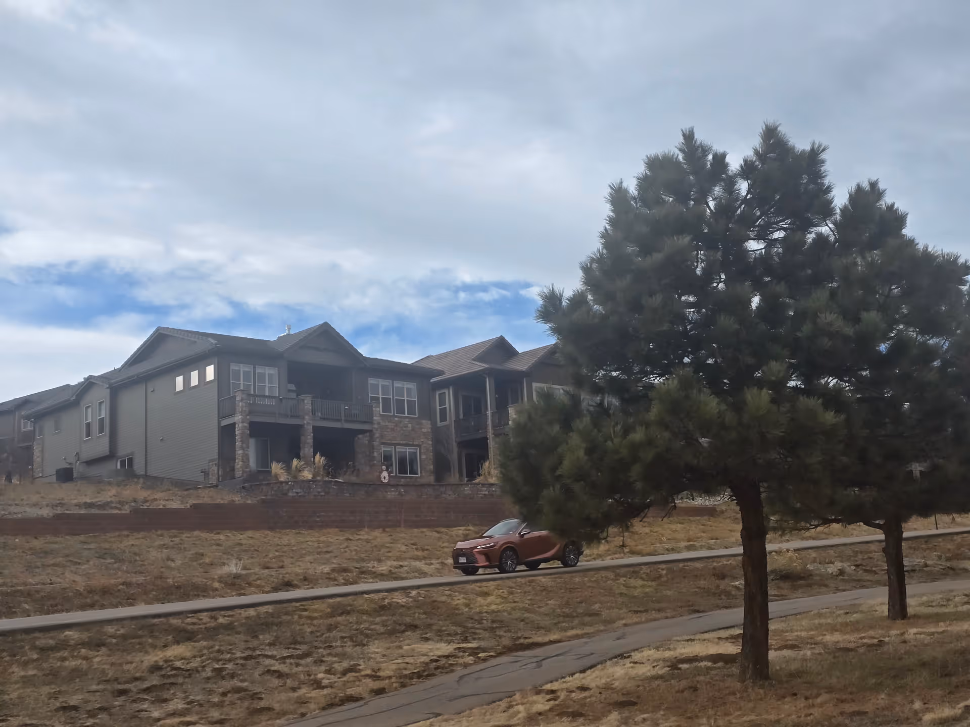 Exterior view of a residential building with stone and siding facade, featuring balconies and multiple windows. In the foreground, there is a paved pathway, a parked car, and two pine trees. The sky is partly cloudy.