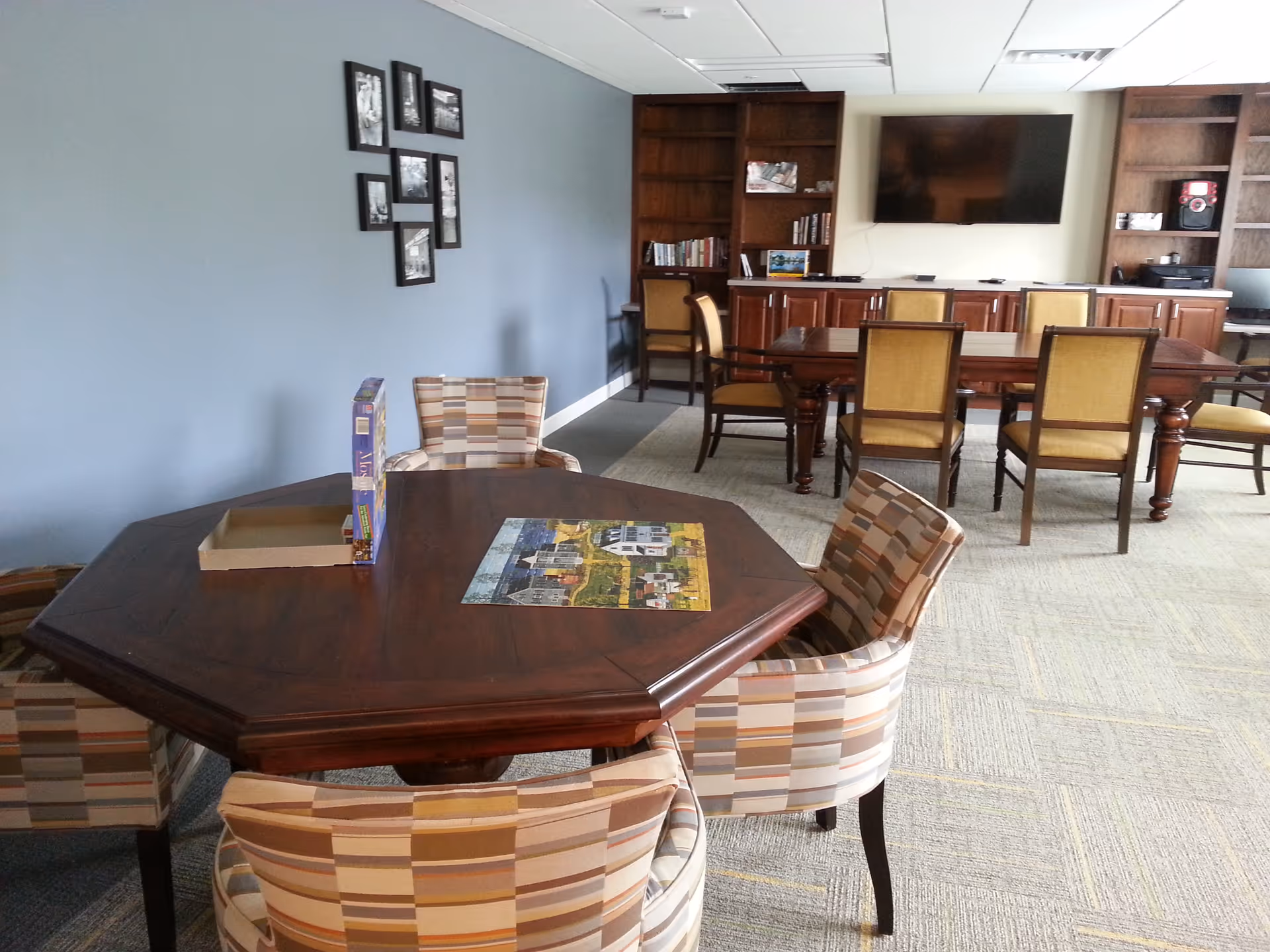 A common area in a senior living facility featuring a wooden octagonal table with a partially completed jigsaw puzzle and a puzzle box on it, surrounded by four patterned upholstered chairs. In the background, there is a rectangular wooden table with six yellow cushioned chairs, built-in wooden shelves with books and decor, and a large flat-screen TV mounted on the wall.