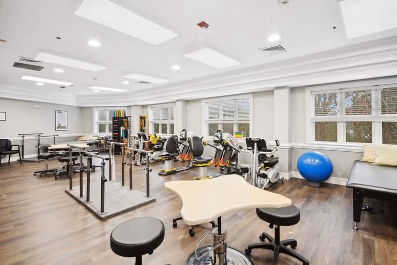 A spacious physical therapy or rehabilitation room with exercise equipment including stationary bikes, parallel bars for walking practice, therapy tables, stools, and a large blue exercise ball. The room has large windows with white blinds, wood flooring, and bright overhead lighting.