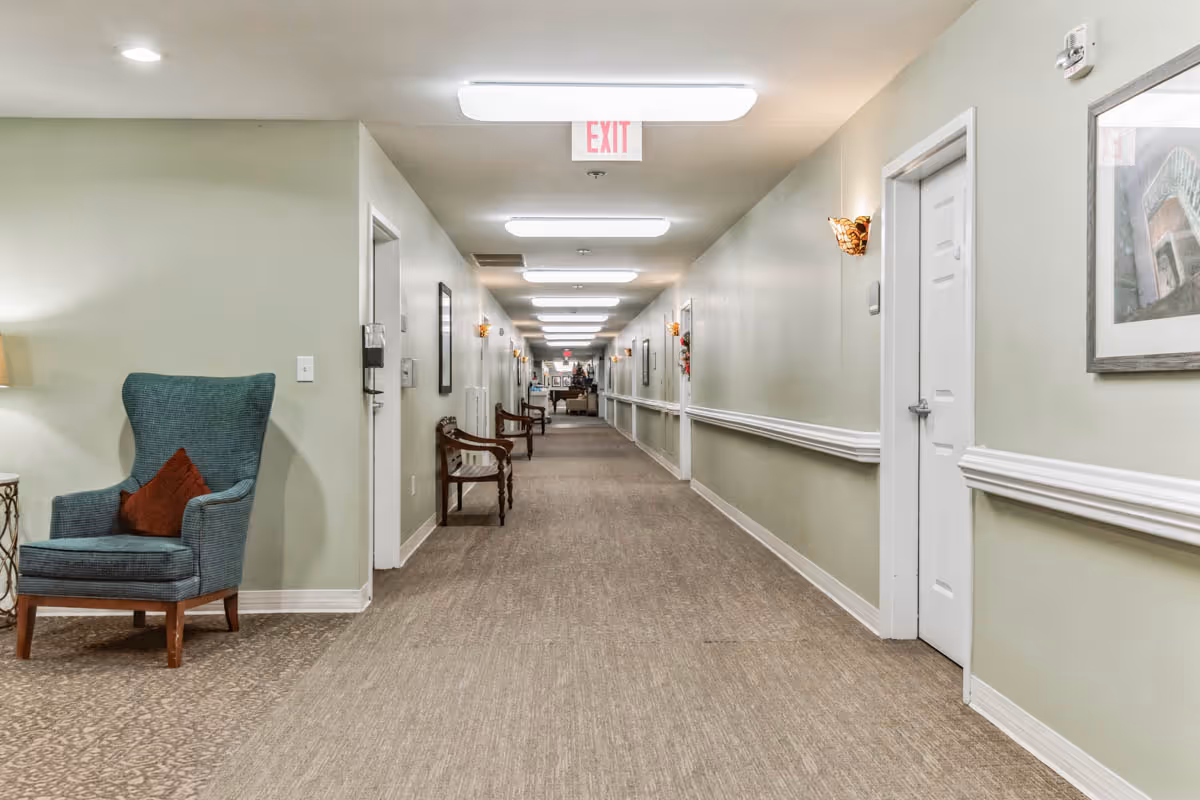 A long, well-lit hallway in a senior living facility with light green walls, beige carpet, and white trim. The hallway features several wooden chairs along the walls, a blue upholstered armchair with a red pillow on the left, framed artwork, and an exit sign on the ceiling at the far end.
