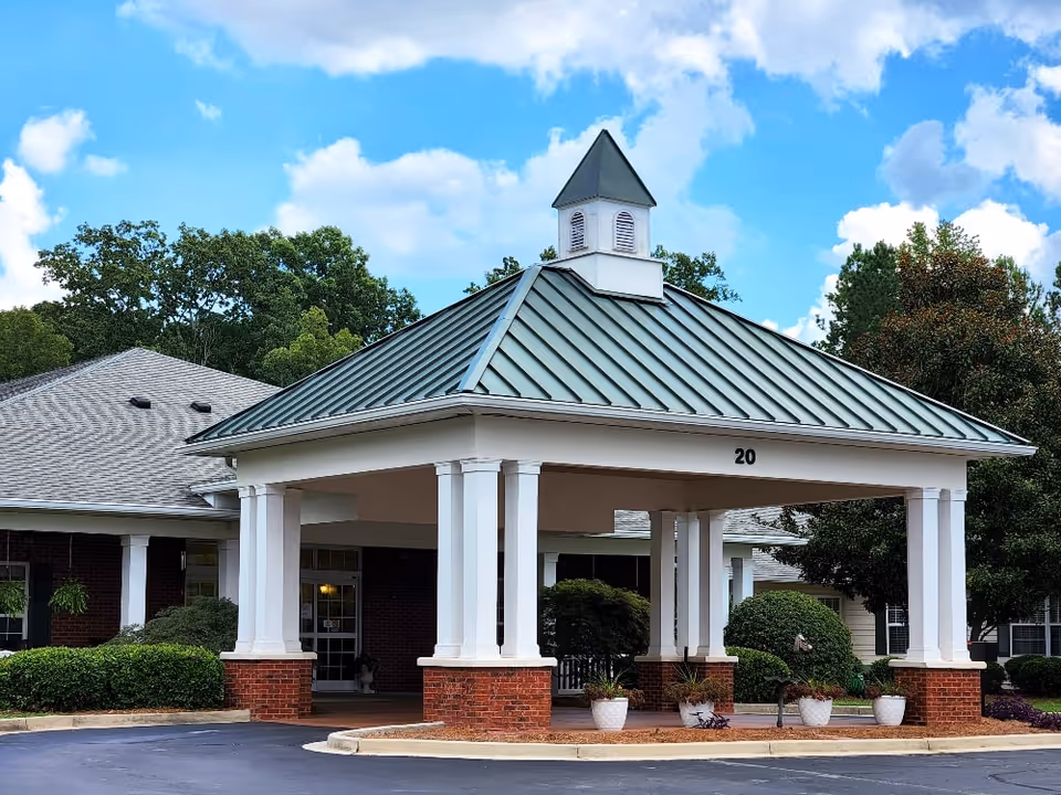 Covered entrance with white columns and a green metal roof above a brick base at the front of a senior living building numbered 20, with shrubs and blue sky.