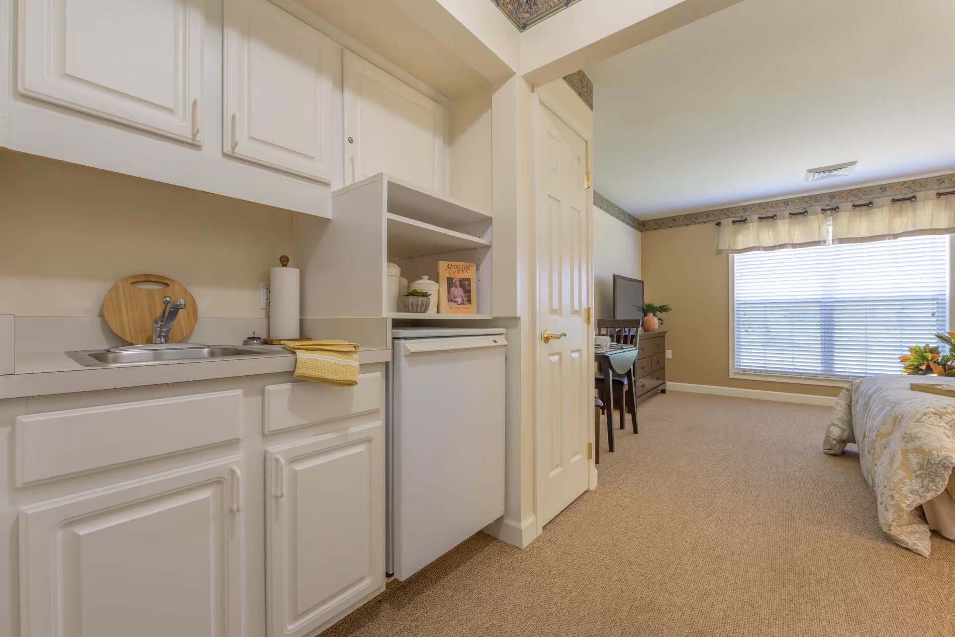 Interior view of a senior living facility room at Brookdale Wooster featuring a small kitchenette with white cabinets, a sink, and a mini refrigerator. The room extends into a bedroom area with a bed, a dresser with a TV on top, a chair, and a large window with blinds and a valance allowing natural light to fill the space.