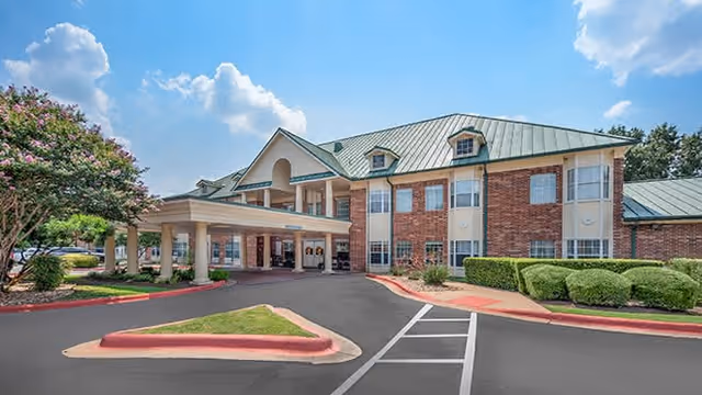 Front entrance of a two-story brick senior living building with a covered porte-cochère, green metal roof and landscaped driveway under a blue sky.