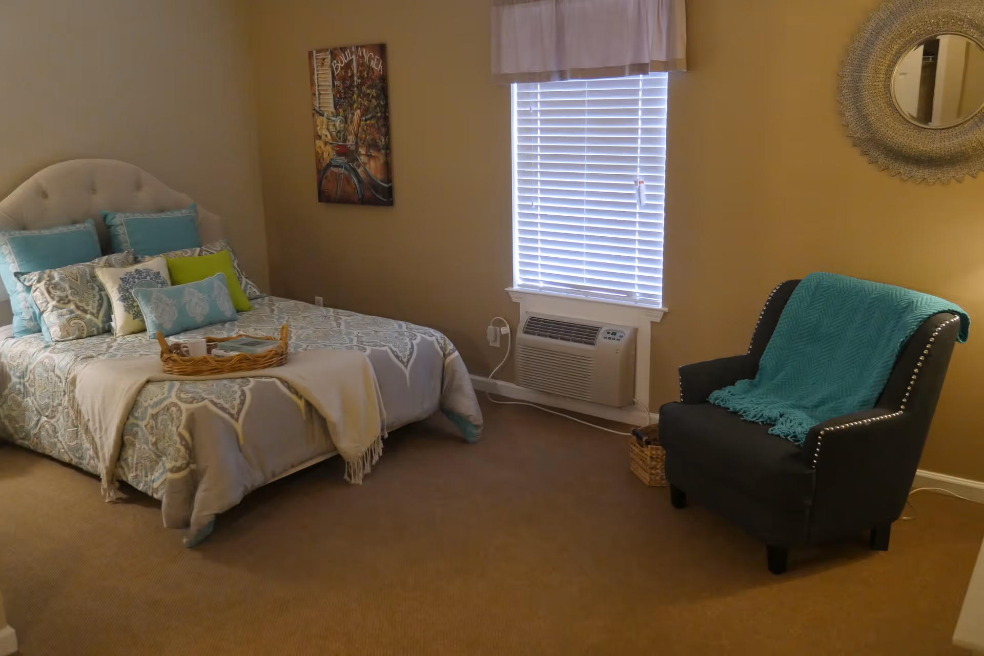 A cozy bedroom with a bed featuring a patterned comforter and multiple pillows in shades of blue and green. A wicker tray with a cup and a book rests on the bed. To the right, there is a dark upholstered armchair with a teal throw blanket draped over it. The room has beige walls, a window with white blinds and a valance, a wall-mounted air conditioning unit below the window, a decorative round mirror on the wall, and a colorful painting hanging beside the bed.