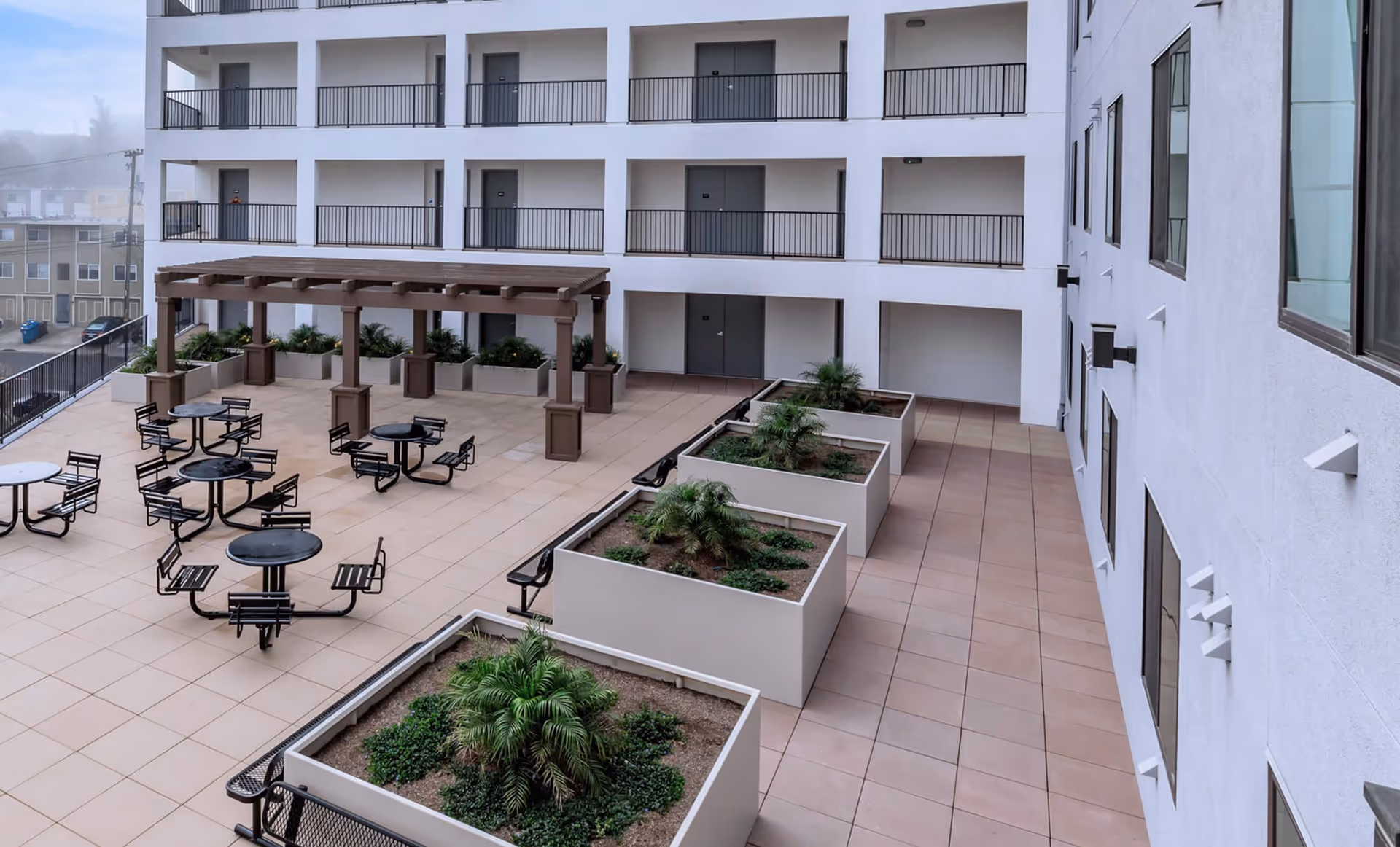 Outdoor courtyard area of Brunswick Street Apartments featuring several round tables with attached chairs, a wooden pergola, and large rectangular planters with green plants. The courtyard is surrounded by a multi-story building with balconies and doors.
