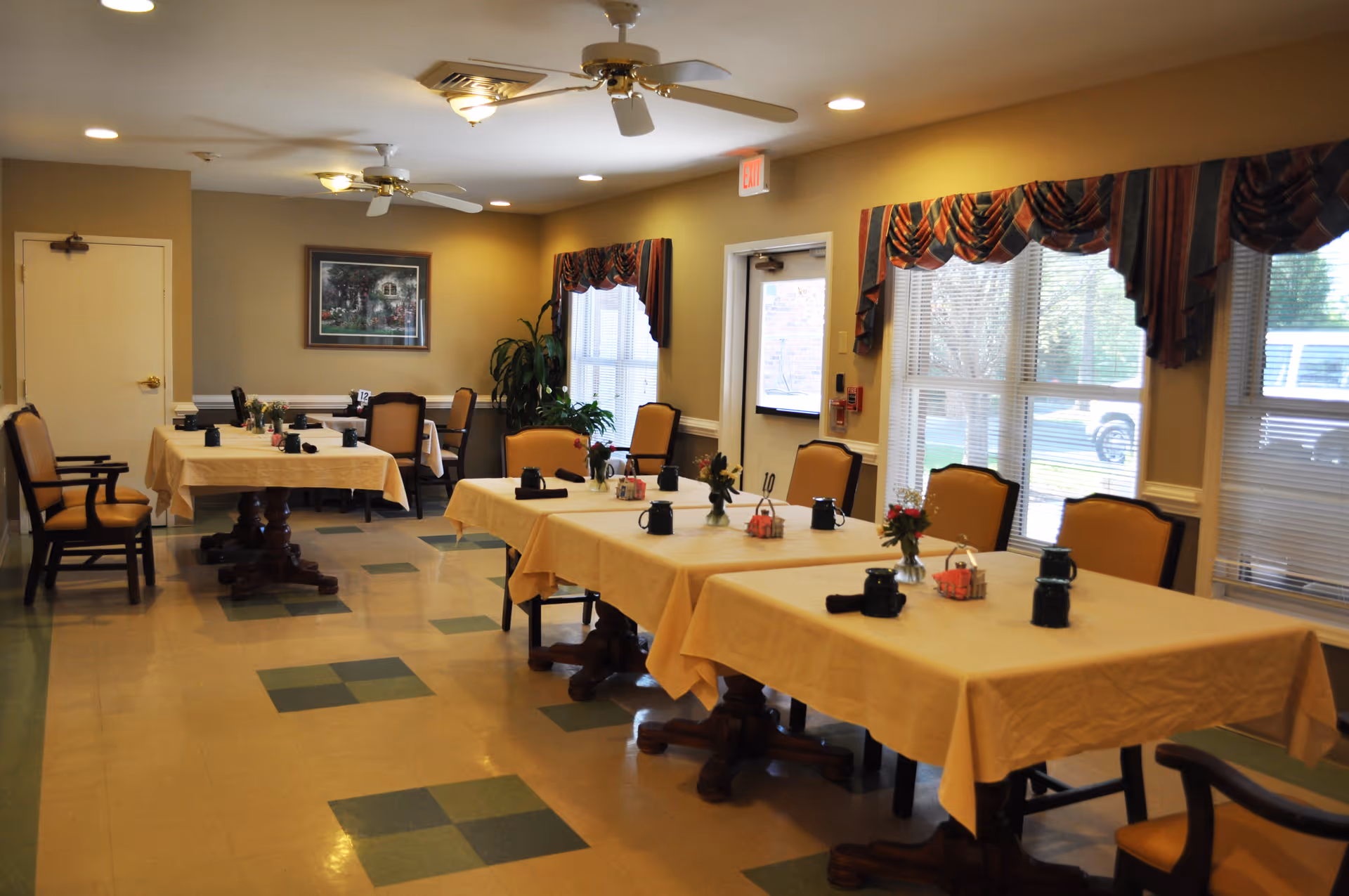 Dining room with multiple tables set with tablecloths, chairs, and small centerpieces beside large windows.