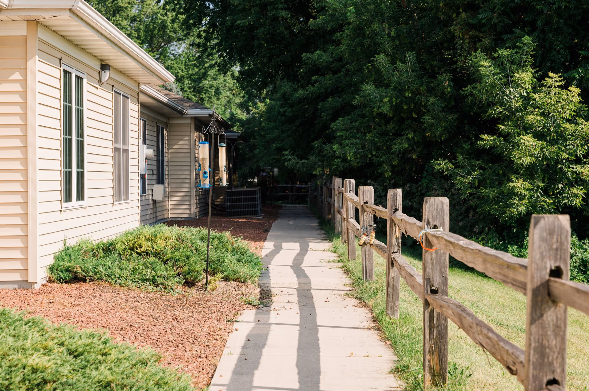 Sidewalk beside a beige siding building with a wooden fence, landscaping, and trees along the path.