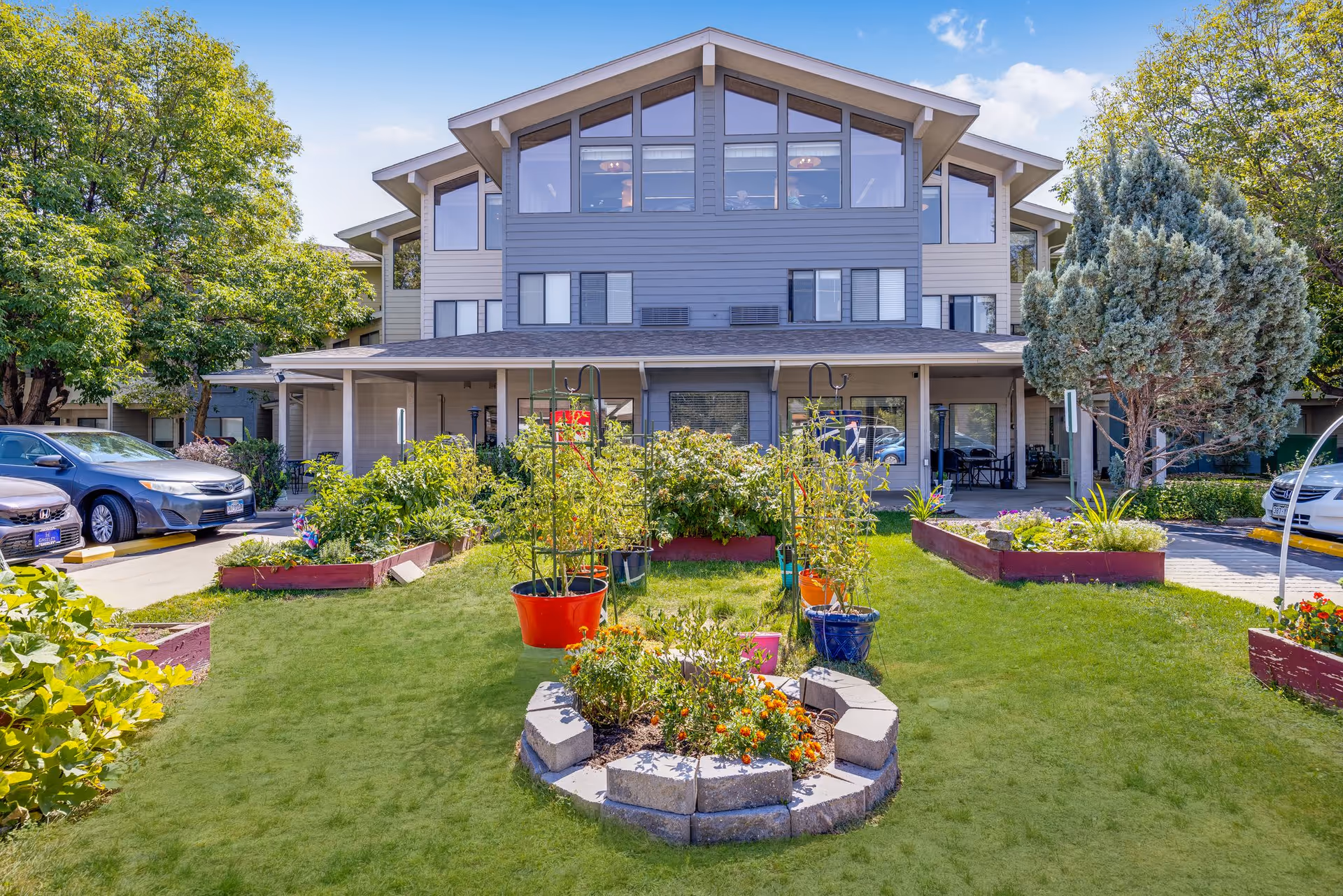 Front exterior view of a multi-story senior living facility named Greeley Place with a well-maintained garden featuring various plants and flowers in pots and raised beds, surrounded by parked cars and trees under a clear blue sky.