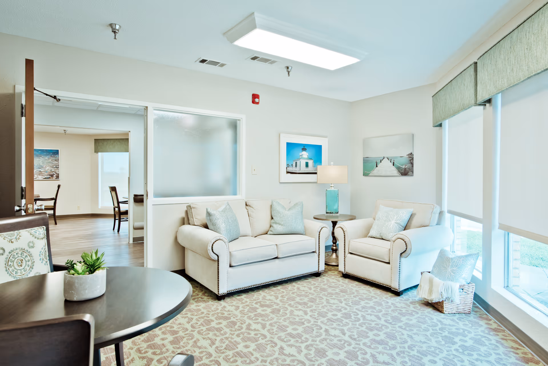 A bright and cozy living room area with a beige loveseat and armchair, both adorned with light blue and patterned throw pillows. A round wooden side table with a turquoise lamp sits between the seating. The room has large windows with white roller shades, a patterned carpet, and two framed pictures on the walls. A round table with a small potted plant is visible in the foreground, and an open doorway leads to another room with chairs and a window.