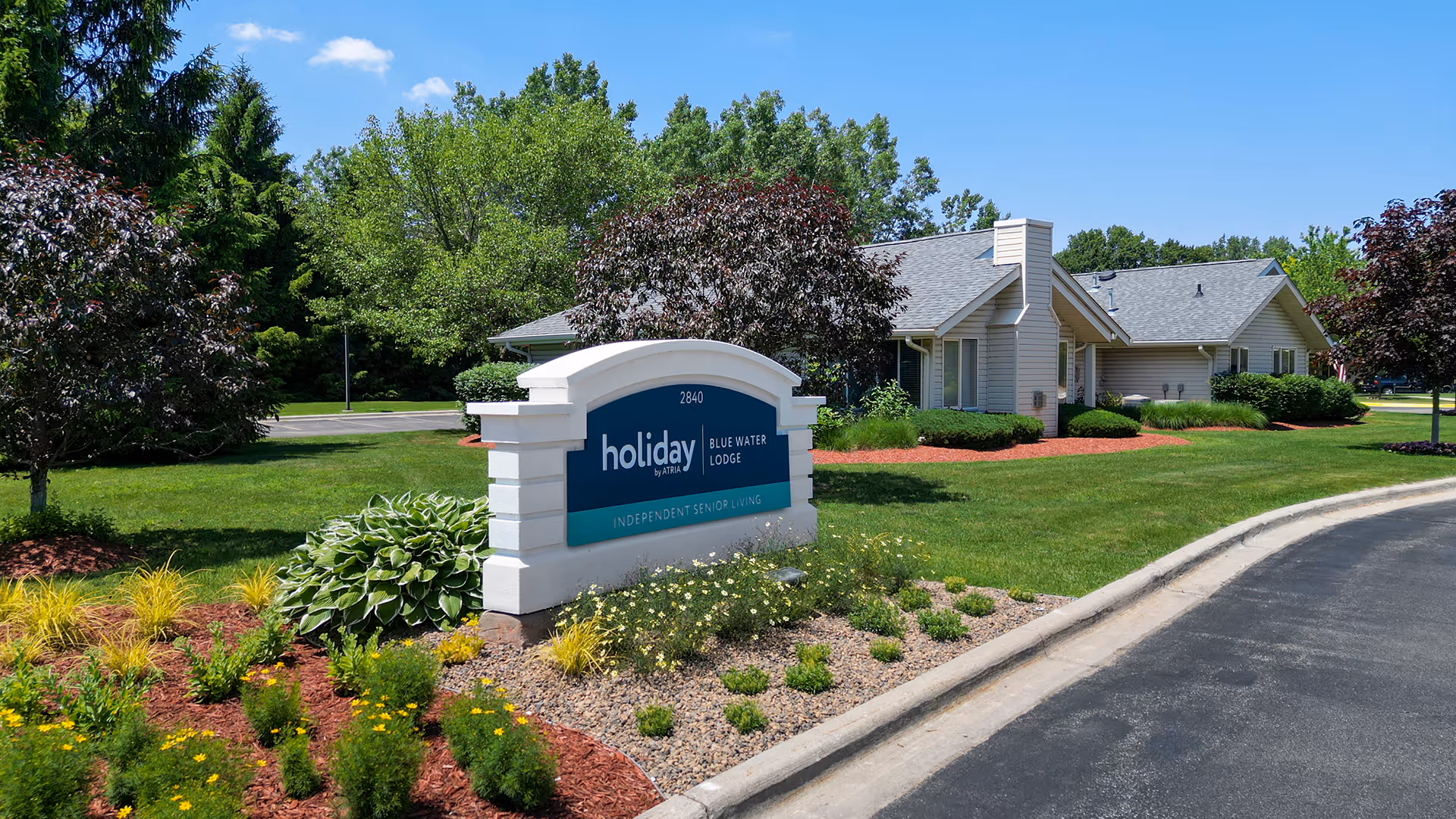 Outdoor view of Holiday Blue Water Lodge senior living facility sign surrounded by landscaped greenery, flowers, and trees with a building and clear blue sky in the background.