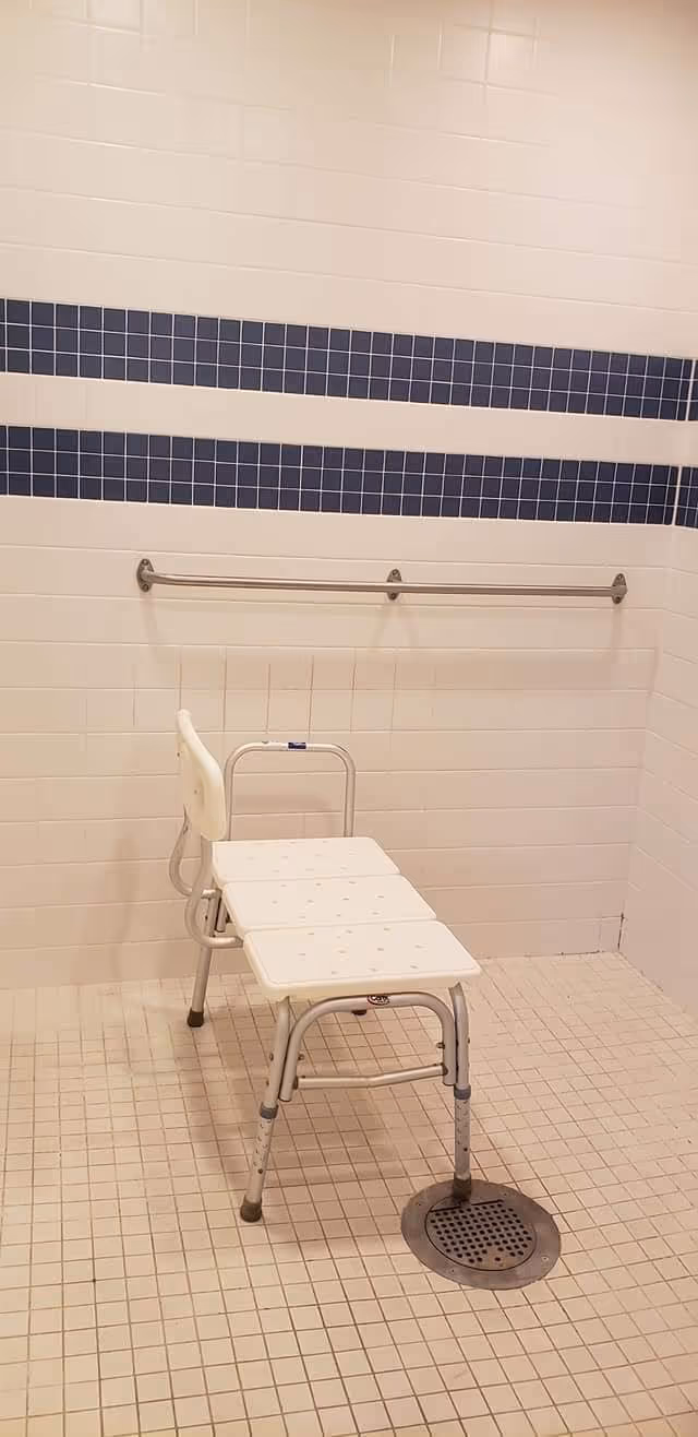 A shower area with white tiled walls and floor, featuring a white shower chair with a backrest and a metal grab bar mounted on the wall with blue tile accents.