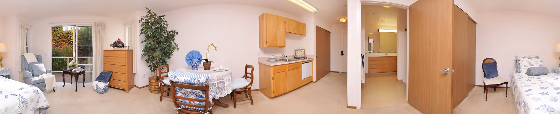 Panoramic studio apartment interior showing a bed, seating area, dining table, small kitchenette, and a bathroom doorway.
