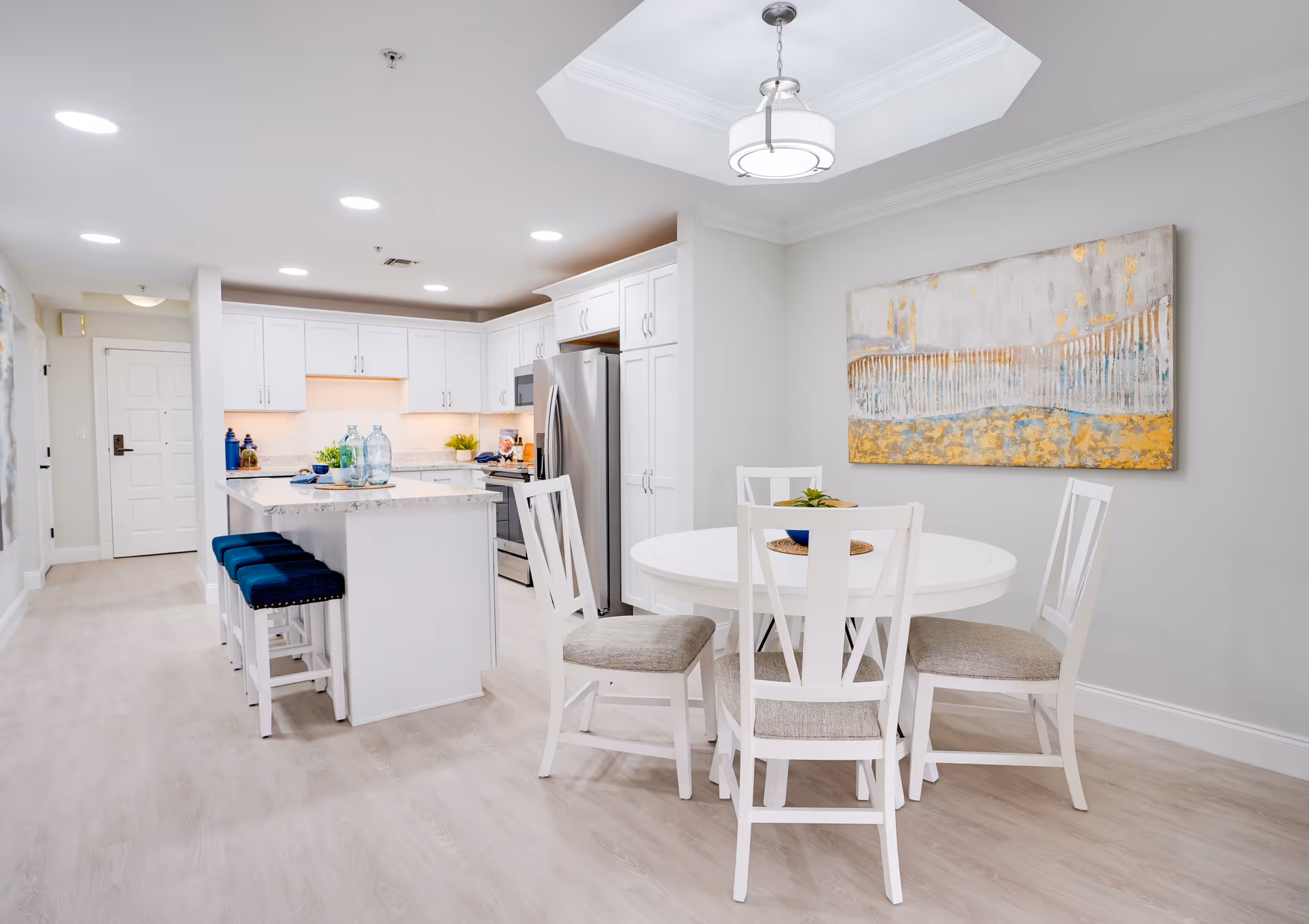 Bright and modern kitchen and dining area with white cabinetry, stainless steel appliances, a kitchen island with three blue cushioned stools, and a round white dining table with four chairs. A large abstract painting with gold and blue tones hangs on the wall above the dining table.