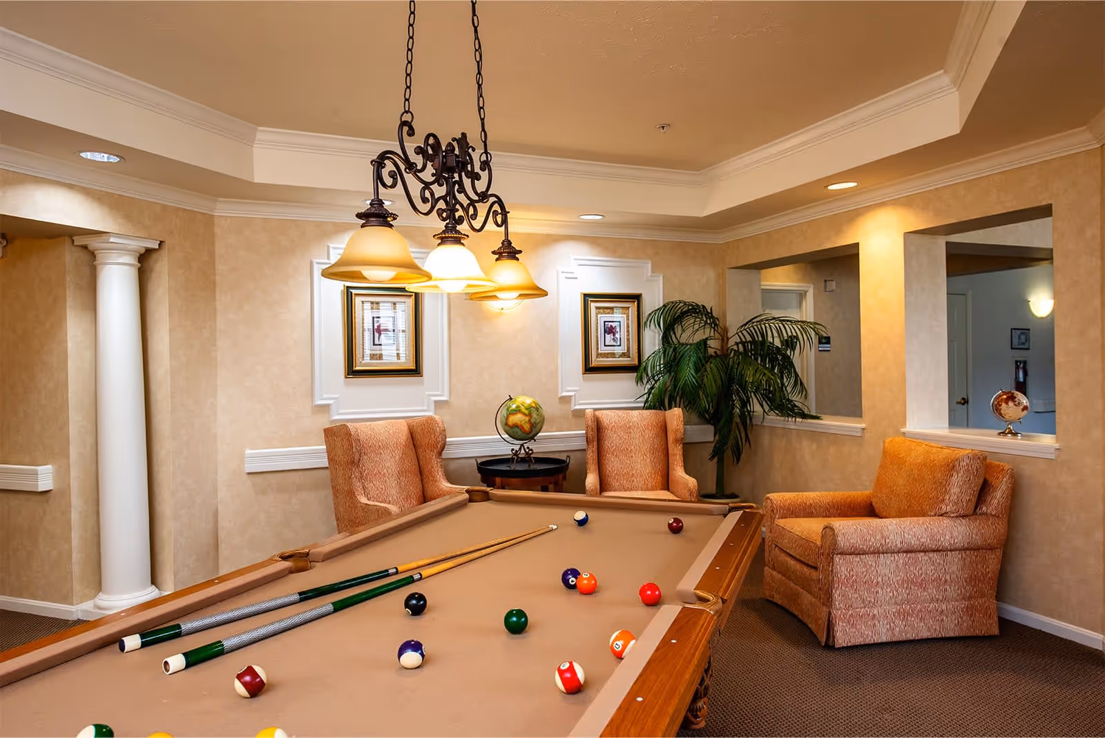 A cozy interior room featuring a pool table with several billiard balls and cues on it. Surrounding the pool table are three upholstered armchairs in a warm, patterned fabric. The walls are beige with white trim and decorative molding, and there are two framed pictures hanging on the wall. A small round table with a globe sits between two of the chairs, and a large potted plant is placed near the corner. A decorative chandelier with four lights hangs above the pool table.