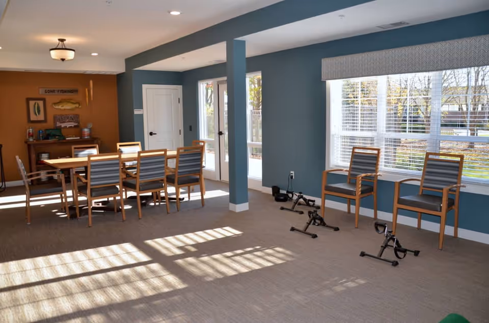 A bright room with blue and brown walls featuring a wooden dining table with eight chairs, two armchairs by a large window, and three pedal exercisers on the floor. The room has large windows letting in natural light and a door leading outside. There is a small wooden cabinet with fishing-themed decorations on the wall above it.