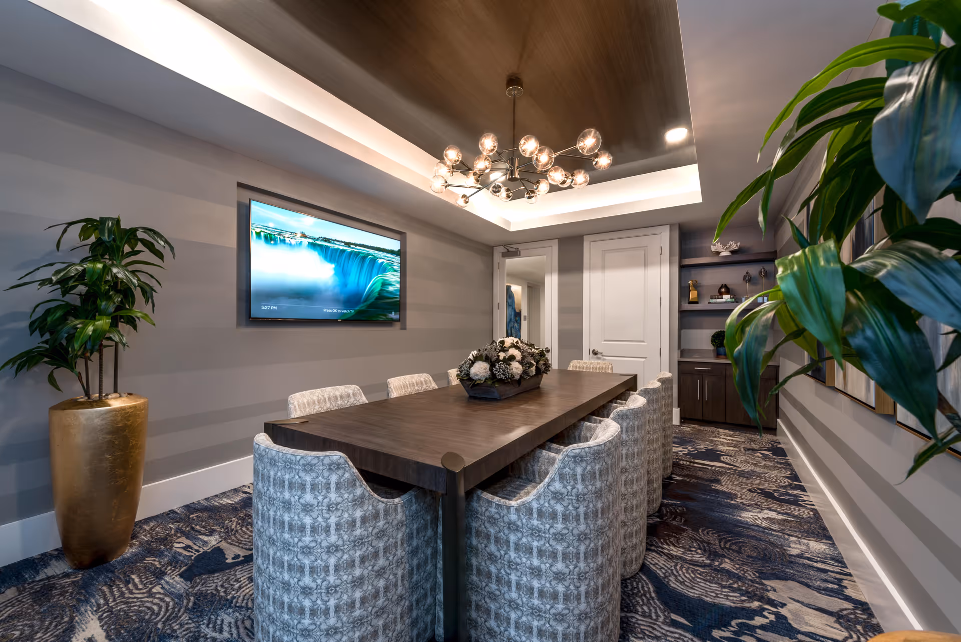 A modern conference or meeting room with a long wooden table surrounded by eight upholstered chairs. The room features a decorative ceiling with recessed lighting and a contemporary chandelier. A large flat-screen TV is mounted on the wall displaying an image of a waterfall. There are two large potted plants on either side of the room, patterned carpet flooring, and built-in shelves with decorative items in the background.