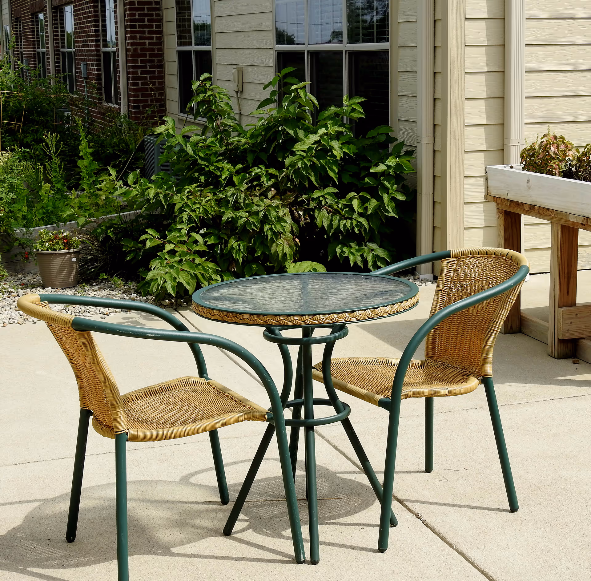 A round glass-topped patio table with three wicker chairs on a concrete patio beside a building and green shrubs.