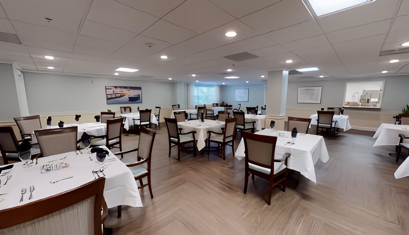 A spacious dining room with multiple tables covered in white tablecloths, each set with black napkins and glassware. The room has wooden chairs with cushioned seats and backs, light-colored walls, and a ceiling with recessed lighting. There is a large window with blinds and a framed picture on the wall.