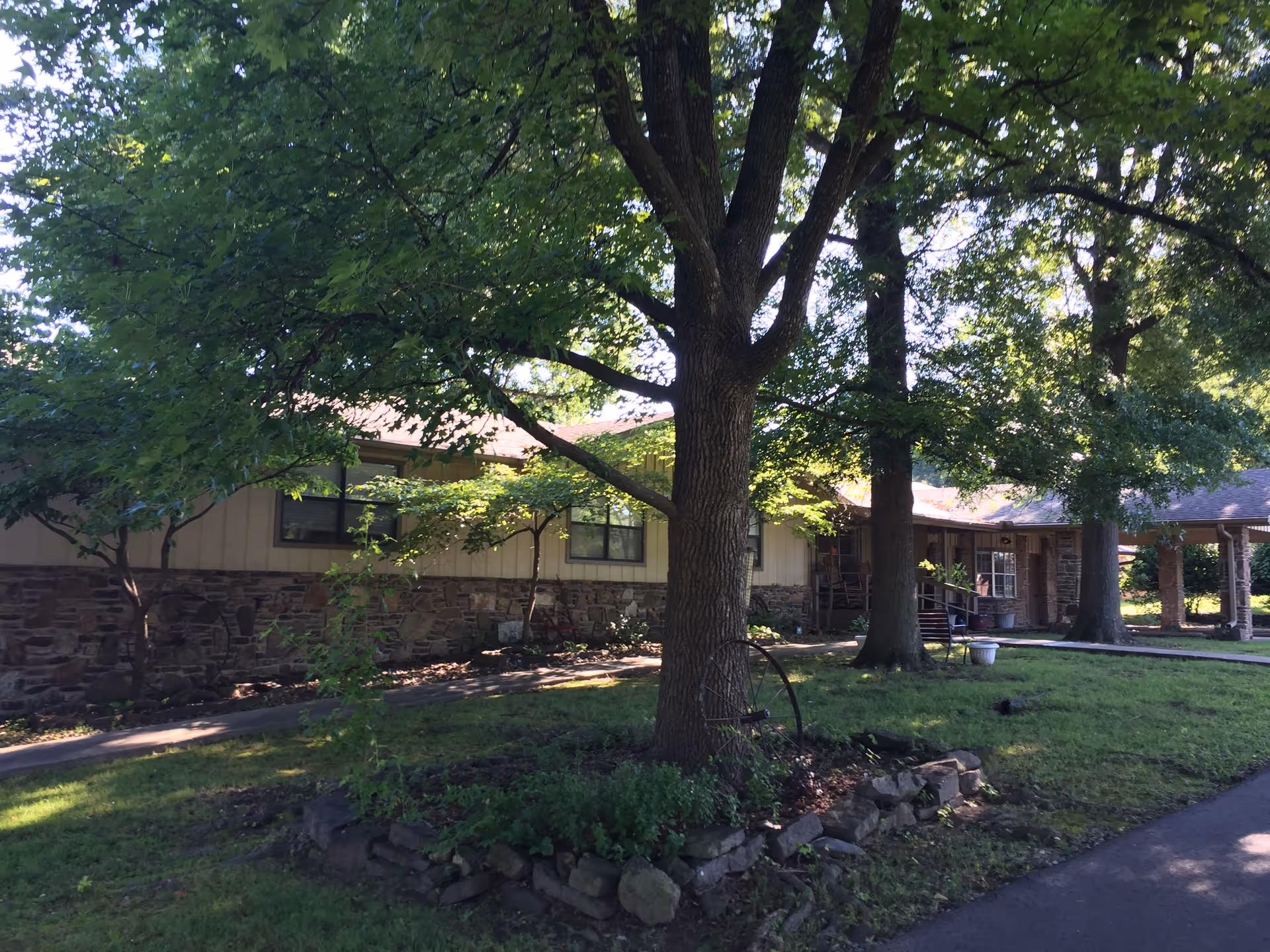 Single-story building front with a stone foundation, large shade trees, lawn, and a covered entrance.